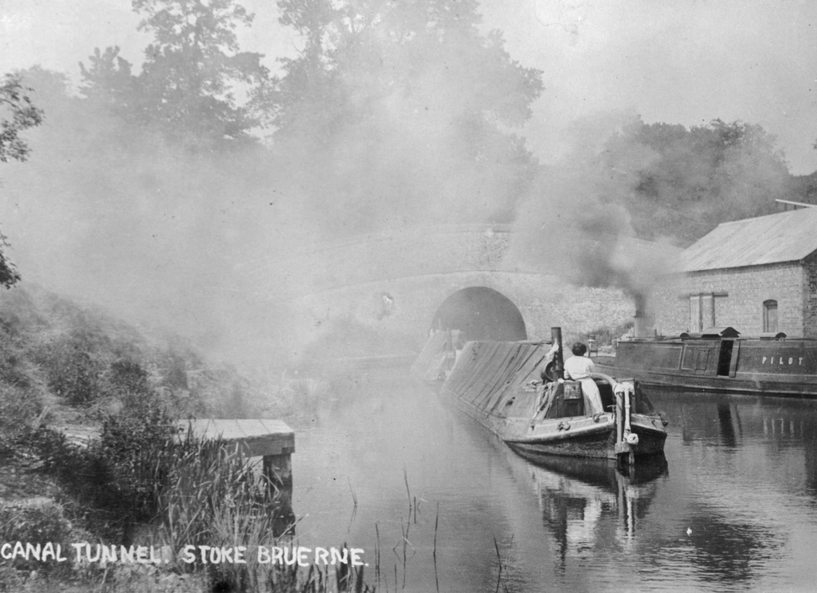An evocative canal scene at the Stoke Bruerne entrance to the tunnel. A pair of buttys (unmotorised boats) are waiting for the tug 'Pilot' (seen at right) to take them through the tunnel. The boats can be seen hanging about mid channel which suggests they expect to be towed through the tunnel quite soon. There is a lot of smoke in the air which is coming from the tug and this is because it was powered by a steam engine! No doubt it was very smoky in the tunnel!